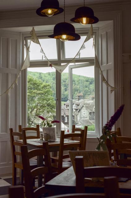 Square wooden tables, surrounded by wooden chairs, a large window with a view to a green fell in the distance and some grey stone houses closer by. Three lightbulbs covered by bowler hats hanging from the ceiling.