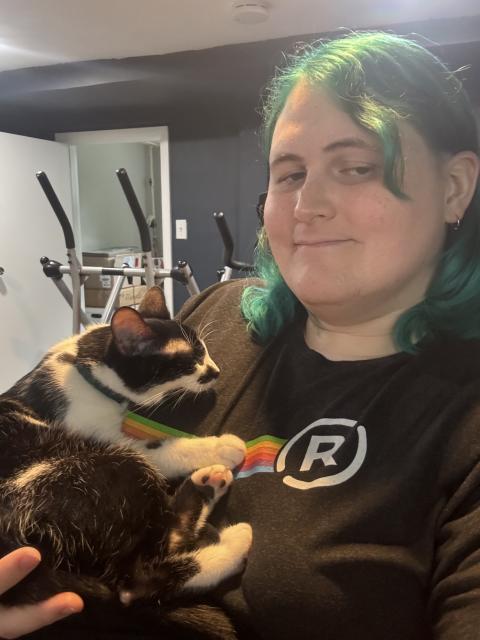 Person with shoulder length teal hair holding a small sleeping tuxedo cat against their chest