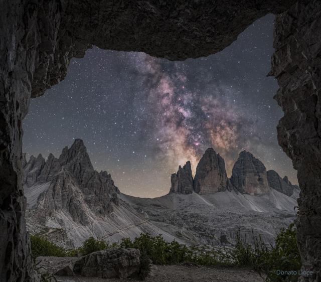 A starry sky is seen through a rectangular rock opening. Three rocky peaks appear toward the right, while other peaks appear toward the left. Rising above the right peaks is the central band of our Milky Way Galaxy.