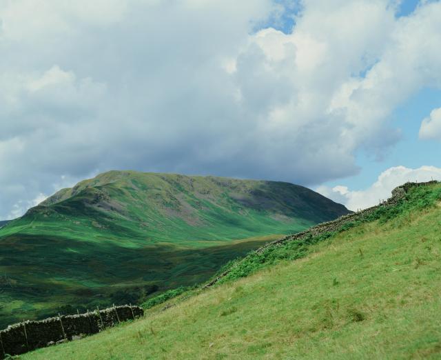 A massive green fell in the distance under a blue sky and white clouds. You can clearly see the shadows of the clouds on the ground.