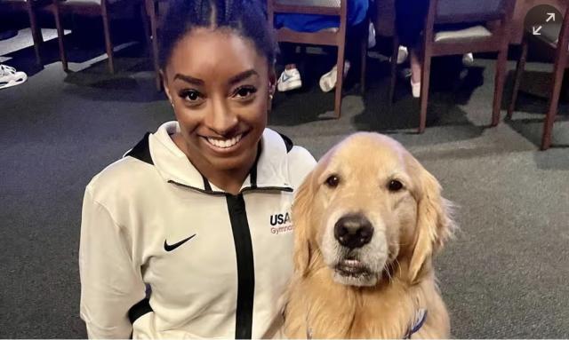 Simone Biles in a white track jacket with a Nike logo smiles at the camera with her arm around a golden retriever also staring at the camera who obviously knows he is the goodest boy