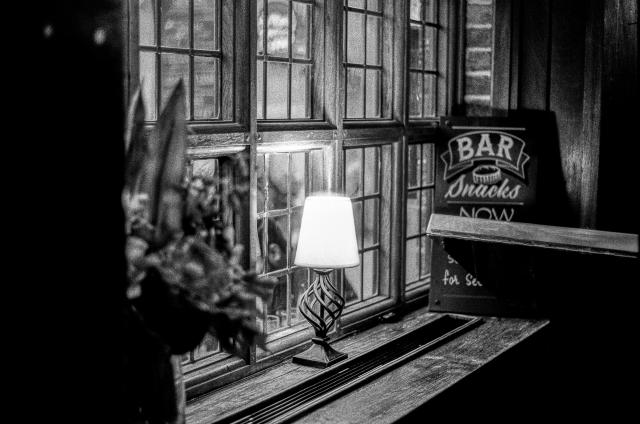 A little table lamp standing on the sill of a multi-lite window. Besides it, there is a sign that reads, over multiple lines, "Bar, snacks, now". The rest of the sign is obstructed by a piece of wood.