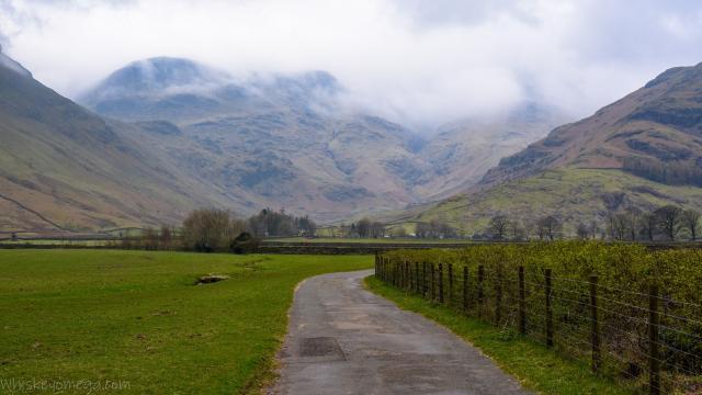 A small mountain in the clouds with slops from hills either side and a farm track in the foreground