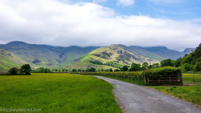 A hill lit up by the sun in front of a ridge of hills in the clouds with a farm track and fields in the foreground with trees