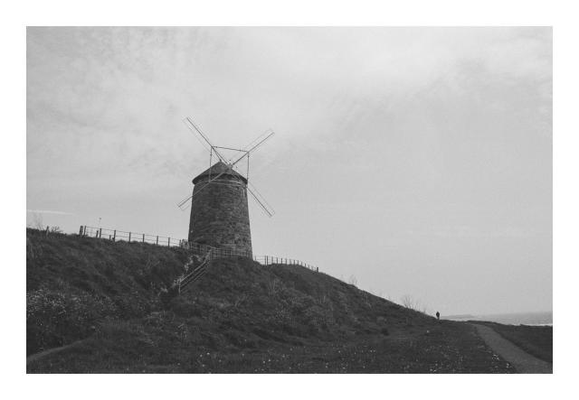 A black and white photograph of a windmill on a low hill, with a path on the right and a figure in the extreme distance. 