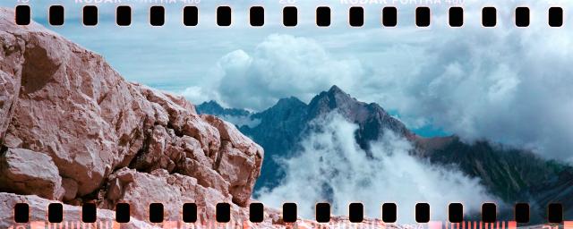 Horizontal landscape panoramic of mountain scenery. Foreground is reddish brown cliff, background is a mountain peak surrounded by white clouds. Film sprocket holes visible on the long edges.