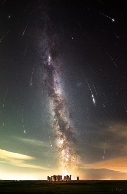 A night sky filled with stars and the band of our Milky Way galaxy is shown also filled with many streaks. The short streaks are all coordinated and together indicate a flow from the top of the image to the bottom. In the foreground at the bottom of the frame is Stonehenge.