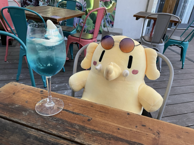 A stuffed Mastodon toy wearing round sunglasses sitting in front of a cocktail glass with a blue drink that has a gummy shark in it. 