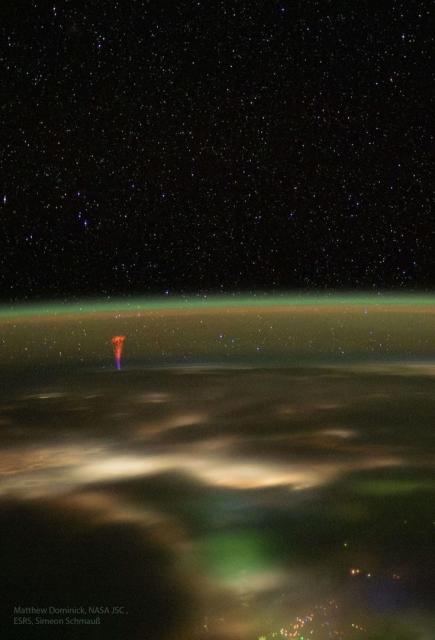 The edge of the Earth is seen from space at night. The curving horizon is tinted green but a red and blue jet rises from left.