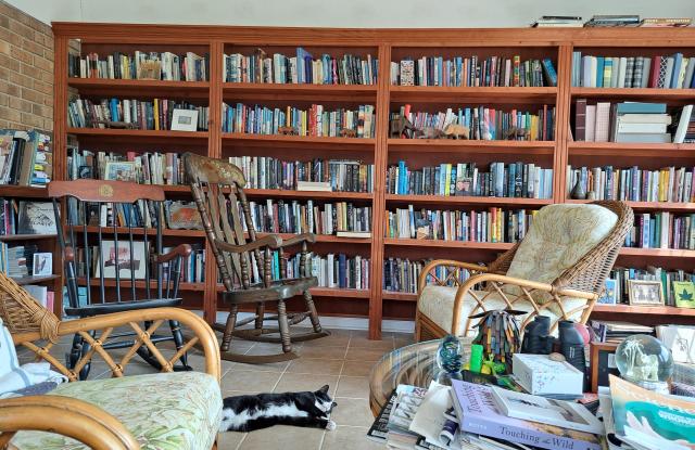 The picture is taken in our room reserved for reading, with floor to near-ceiling cases stuffed with books and rattan and wood chairs thatcrock. Marie, a tuxedo cat, lays sprawled on the floor. There is some clutter on a glass table filled with books, magazines, and papers, but she ignores this as do I. 