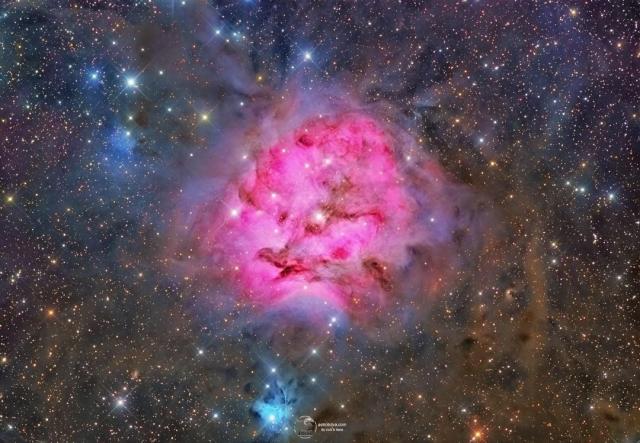 A red emission nebula is shown against a busy starfield with many dark dust filaments near the nebula's center. Near the bottom of the image is a smaller blue reflection nebula.