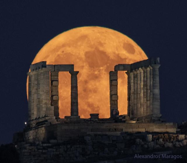 A large and orange-tinted moon is pictured rising beyond the pillars of an ancient structure. The foreground is dark and the night sky behind the Moon appear blue.