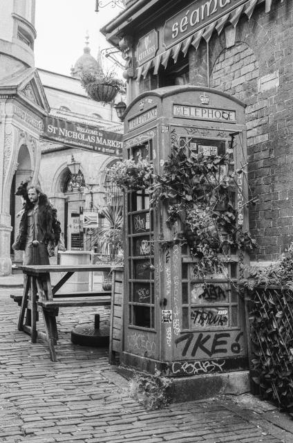 An old telephone box overgrown by plants and covered in tags standing next to a pub. In the background, a banner across the street welcoming passersby to the St. Nicholas Market. A man with a cigarette in his mouth is walking past, with his dog wrapped around his neck like a fancy scarf.