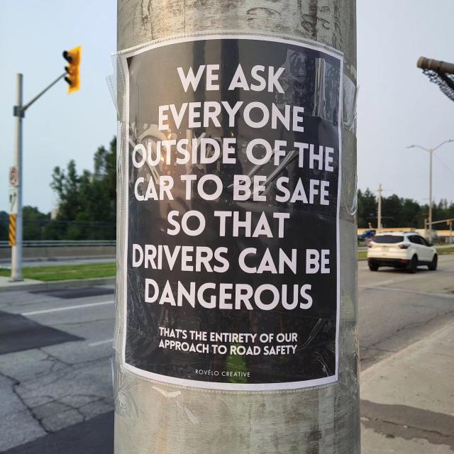 A roadside poster taped to a lamppost.
It reads, “We ask everyone outside of the car to be safe so that drivers can be dangerous.”. And in smaller typeface at the bottom, “That’s the entirety of our approach to road safety.”