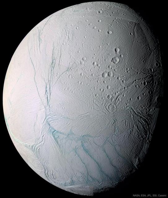 A light-colored spherical body is shown mostly illuminated against a dark background. Many craters are visible. Unusual blue stripes meander on the surface.