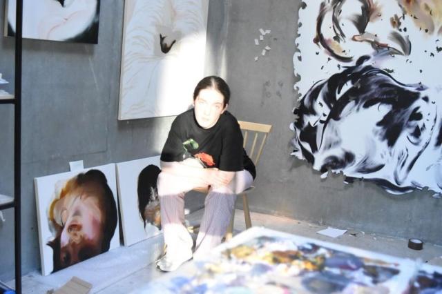 Image: colour photograph: we see a figure sitting on a wooden chair, leaning forward, and caught in bright sunshine from a window; it is probably the artist; on the walls are artworks of various sorts, possibly in progress, and there is a hint of a horizontal artwork, possibly on a table, in the foreground; the figure is wearing a black top and pale jeans / trousers, and maybe runners • “Ormston House is excited to announce Sian Costello’s imminent solo exhibition, entitled Hot Child. In this show, featuring several large new works, Costello continues her exploration of representing the female body via performative self-portraiture. The exhibition will open Thursday, 25 July 2024 from 7–9pm.    Those already familiar with this artist’s works will note numerous key developments in the new paintings shown here. Crucially, we see a dissolution of the depicted subject via the presence of more negative space; recognisable representational details are married with gestural abstraction.…”