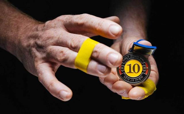 Image: photo or video still of two hands, one holding a medal; the medal has the number 10 in the middle, on a strong yellow, circular background; the text in a band around this circle reads ‘ONE STEP AT A TIME | CÉIM AR CHÉIM’; the background is black; there is bright yellow tape binding fingers on either hand – the middle and ring finger on the right hand, the index and middle on the left hand • “Overcoming addiction is a slow and personal process, involving all our strengths and vulnerabilities. Relapse is not a failure, but a necessary part of the long journey towards change.  During a series of conversations with the Drug & Alcohol Task Force in Tallaght, it became clear that people needed a physical symbol for this journey, to visualise and hold in their hand the path they’ve travelled. These tokens represent victorious moments: 10 years, 5 years, 1 year, 6 months, and perhaps most importantly, the first day—the day of decision and action ("Take the risk or lose the chance")…”