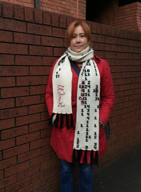 A woman in a red coat in front of a brick wall. She’s wearing a thick white scarf that has embroidered numbers in black and a cursive red Wow!