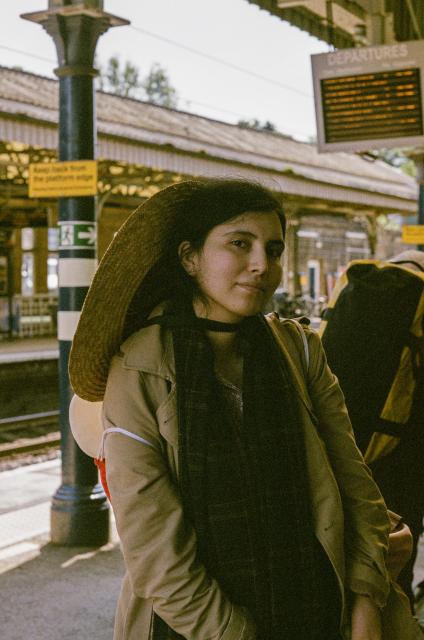 A beautiful woman with black hair, wearing a choker, a big straw hat, a scarf, and a trenchcoat, standing on a train platform, with a pillar and an electronic display of departure times behind her.