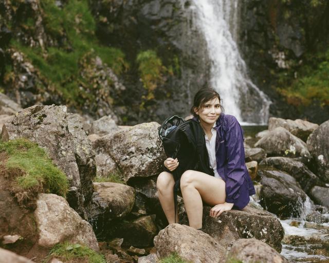 A woman with dark hair, wearing a white shirt and a purple wind jacket, is holding a backpack and glasses while sitting on a rock in front of a waterfall.