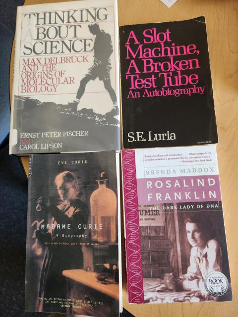 Four books on a table: Max Delbruck and the origins of molecular biology (Fischer and Lipson), A slot machine a broken test tube (Luria), madame curie (Eve Curie), the dark lady of DNA (Maddox)