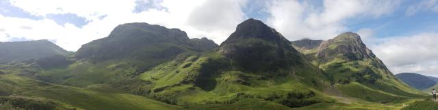 Eine Panoramalandschaft mit mehreren großen Bergen, "The Three Sisters" in Glencoe, die sich unter einem teilweise bewölkten Himmel erstrecken. Die Berge, mit teils steilen und felsigen Hängen, sind mit saftigem Grün überzogen, das von den Tälern bis zu den höheren Gipfeln reicht. Die Berge stehen in einer Reihe und dominieren das Bild. Im Vordergrund erstreckt sich eine sanft hügelige Wiese, ebenfalls mit Gras bedeckt, die einen Übergang zu den massiven Bergformationen bildet. Der Himmel ist überwiegend blau, mit einigen großen, weißen Wolkenfeldern, die sich über die Szenerie ziehen.