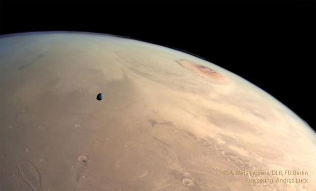 A panoramic view of the surface of Mars. Several landforms are visible including craters and volcanos. A small dark moon is superposed in front of part of the surface.