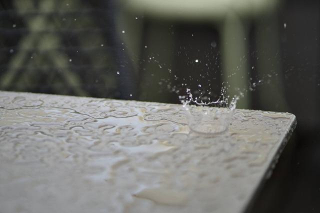 Eine Tischplatte im Regen. Nahaufnahme mit vielen kleinen Pfützen. Ein Tropfen fällt in eine der Pfützen und verursacht einen kreisförmigen Spritzer.