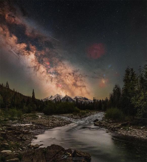 A starry sky is shown with the busy central band of our Milky Way Galaxy showing diagonally from the upper left. Mountains are on the horizon, with trees and a stream running up from the foreground.