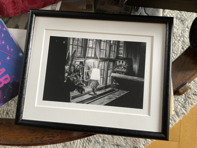 A printed photo in a frame with a passepartout. The photo is black and white and features a lamp in front of a window. 