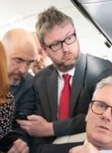 A crop of the same image showing two white male-presenting people in the background. It could be interpreted many ways, but to me one looks like a disgruntled civil servant who is waving an intro to the other, who looks like an angry hitman about to strike.