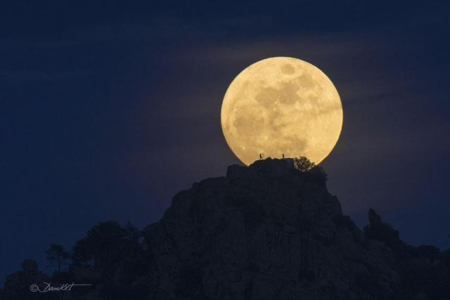 Earth's Moon is shown just beyond a rocky hill. The Moon is near full phase. On the hill the silhouette of a person looking through a telescope can be seen. A rollover darkens part of the Moon that looks to some like a human face.