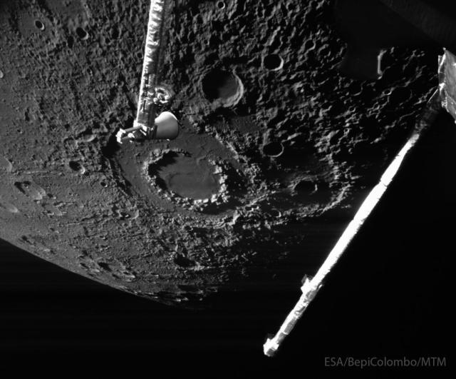 The cratered surface of a large body is shown: Mercury. The largest feature visible is a large impact crater with two rings, near the image center. Arms from the BepiColumbo spacecraft that took the image are seen extending into the image from the top and the right.