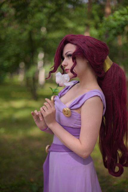 A portrait of a young woman wearing light-lilac-colored dress without sleeves. She has long dark-magenta hair gathered in a large ponytail. She turned to the camera by her left side. Her hands is at her chest level, she holds a white rose in both her hands. She looks thoughtfully with her eyes turned up and left.