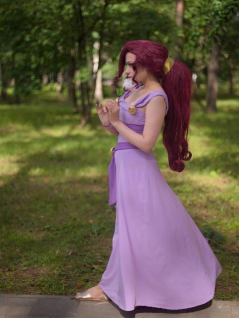 A portrait of a young woman wearing light-lilac-colored dress without sleeves. She has long dark-magenta hair gathered in a large ponytail. She turned to the camera by her left side. Her hands is at her chest level, she holds a white rose in both her hands. She looks pensively forward and down.