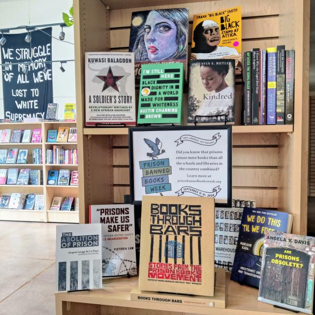 A photo taken inside of Firestorm Books shows a book display for Prison Banned Books Week. A sign on the display reads "Did you know that prisons censor more books than all the schools and libraries in the country combined? Learn more at prisonbannedbooksweek.org." In the background, a banner reads "We struggle in memory of all we've lost to white supremacy and fascism."