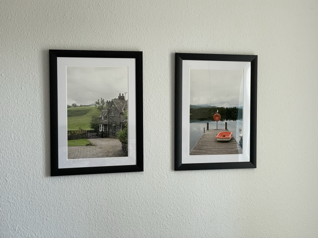 Two framed color prints on a white wall. The left features a stone house in front of a green hill, the right features a seagull standing atop a pier. 