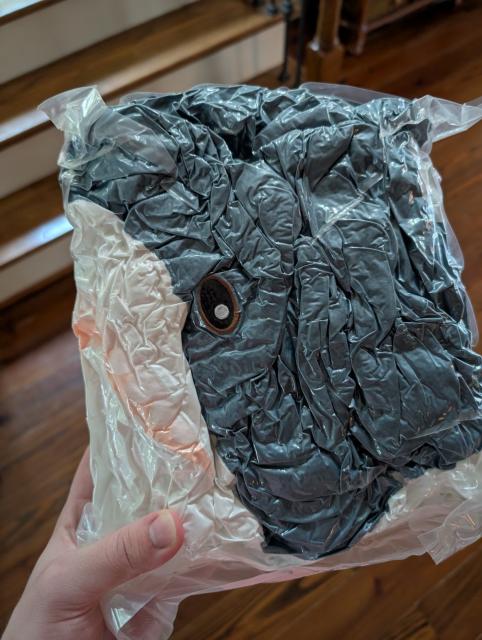 a hand holds a vacuum sealed bag. inside the bag is a blue and white plush shark, a blahaj, with a big eye and a white pupil. the shark is squished and looks very compressed.