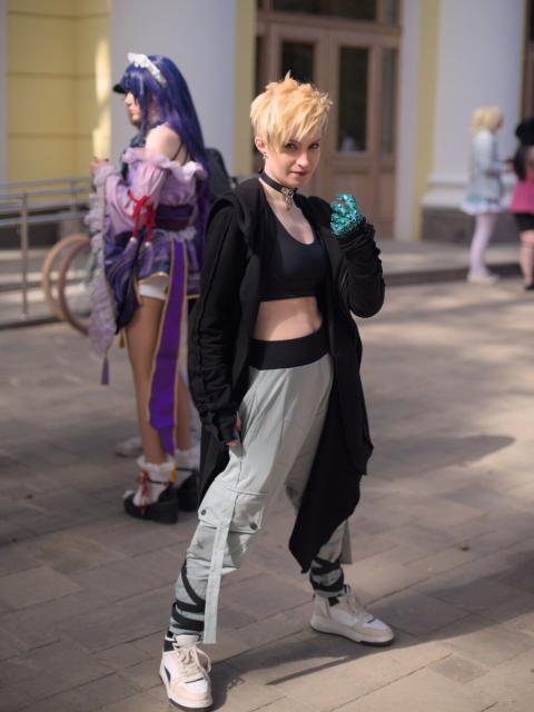 A portrait of a young woman wearing black choker, black tank-top, long black cardigan with a hood, cargo-pants (almost white with light-blue tint) and white sports shoes. She has blue eyes and short yellow-white hair and shaved back of the head. Her left hand raised at a chest level, instead of a palm there is some sort of a prosthetic made of metallic-looking blueish-green plastic.