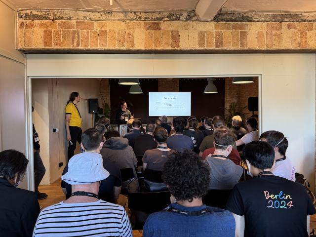 A speaker next to their slides, presenting in front of a very crowded room