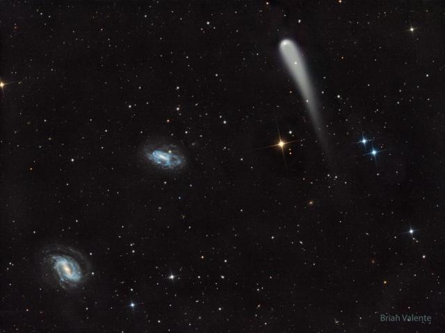 A picture shows a starfield with three prominent objects. A blue spiral galaxy is on the lower left and another blue spiral is just left of center. Toward the upper right is a light-colored comet with a tail fading toward in the image bottom.