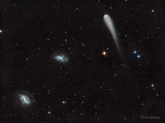 A picture shows a starfield with three prominent objects. A blue spiral galaxy is on the lower left and another blue spiral is just left of center. Toward the upper right is a light-colored comet with a tail fading toward in the image bottom.