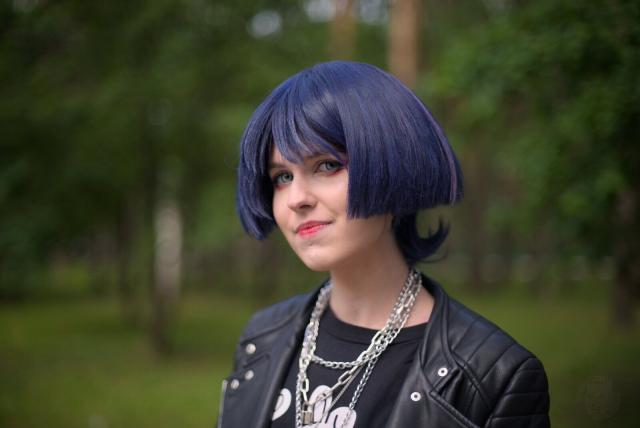 A head and shoulders portrait of young woman wearing black t-shirt with white print, black leather jacket and several necklaces in a form of chains. Her dark-blue hair styled in a short bob. Her eyes are blue. She looks to the camera and smiles only with her eyes.