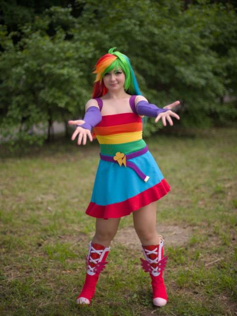 A portrait of a young woman wearing rainbow-striped short dress, purple arms sleeves and soft red knee-length boots. Her hair is rainbow-colored. Her arms are stretched forward with palms up. She looks into the camera and smiles.