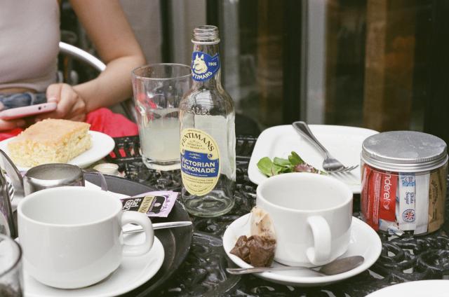 Tea mugs, cake, empty bottle of lemonade next to a glass of lemonade, a used tea bag, a plate with some greens on it, on top of a black metal table. Sitting across, a woman (my wife) holding a pink phone.