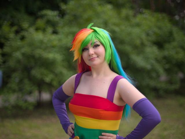 A wais-height portrait of a young woman wearing rainbow-striped short dress and purple arm sleeves. Her hair is rainbow-colored. Her hands are placed on her hips. She looks into the camera and smiles.