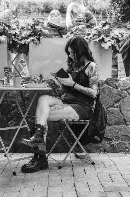 A black and white photo of a woman with long black hair, wearing shorts, a tanktop, and short boots, with tattoos on her arms and legs, sitting cross-legged in front of a cafe table and reading something on the phone.