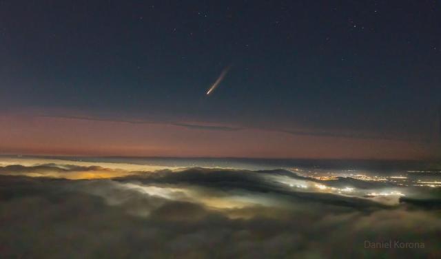 A picture shows clouds across the bottom and a dark night sky across the top. In the middle is a band of orange sky. City lights are visible on the right through gaps in the clouds. In the center of the upper sky is a comet with its tail pointing toward the upper right.