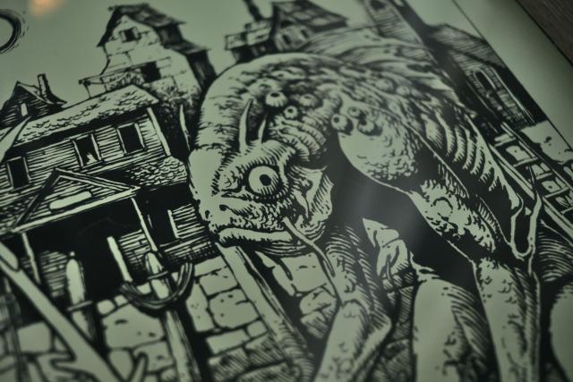 close up of the print showing details of the fish creature covered in mussels with dilapidated buildings in the background