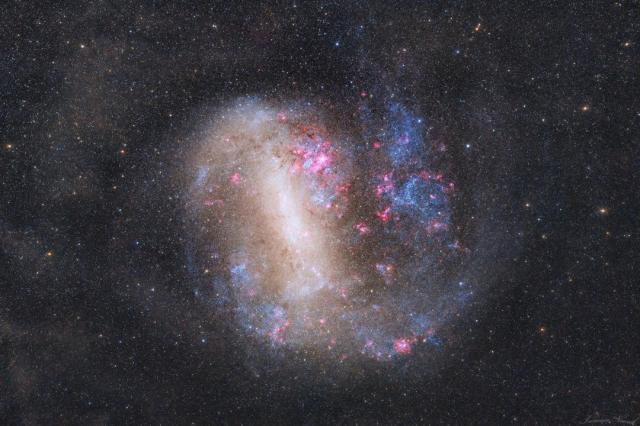 An unusual looking galaxy is shown with a light bar running nearly vertical and blue stars and red nebulas around the edges.